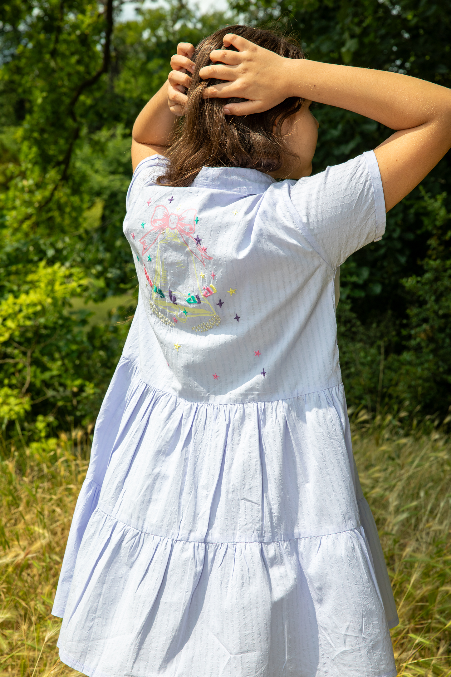 Woman wearing light blue tiered dress with colorful embroidery on back standing outdoors