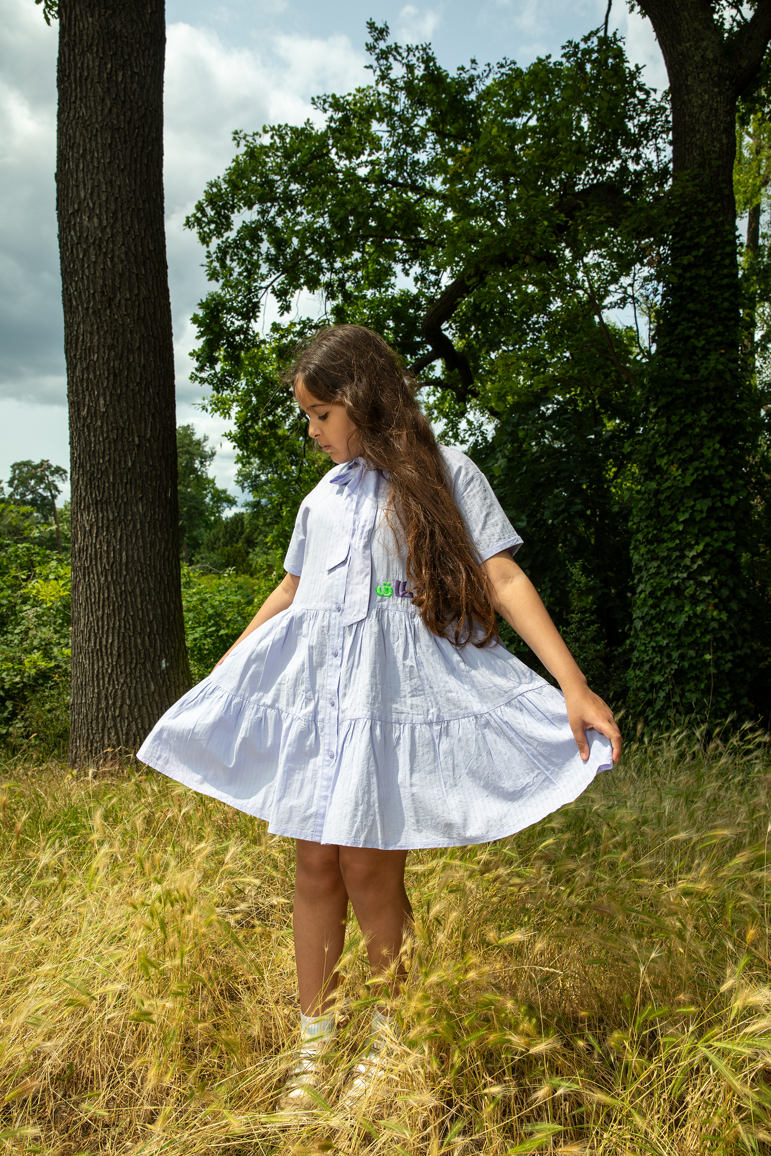 Girl wearing a white short-sleeve dress standing in a grassy field surrounded by trees