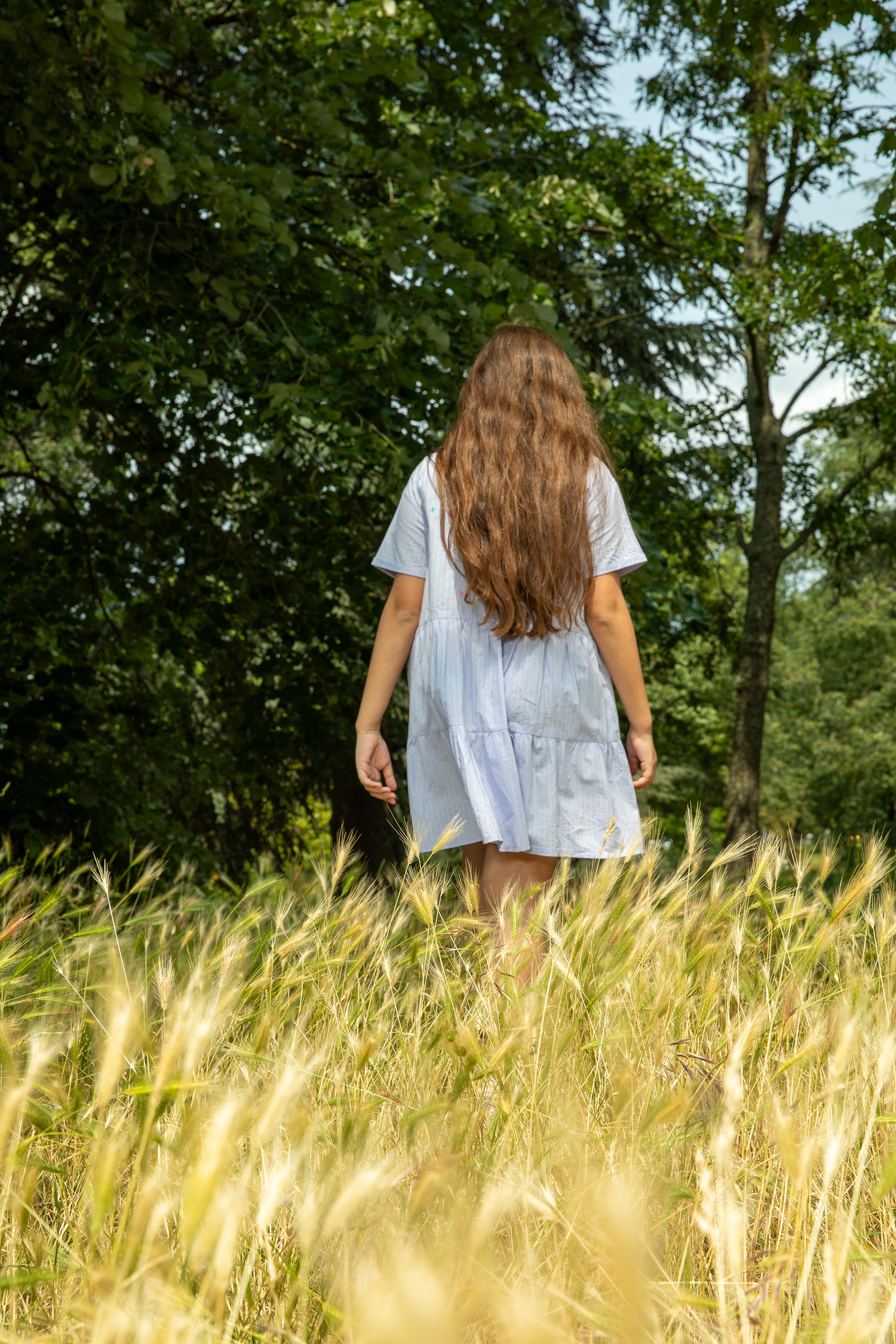 Woman with long hair wearing a white summer dress walking through a grassy field surrounded by tall trees