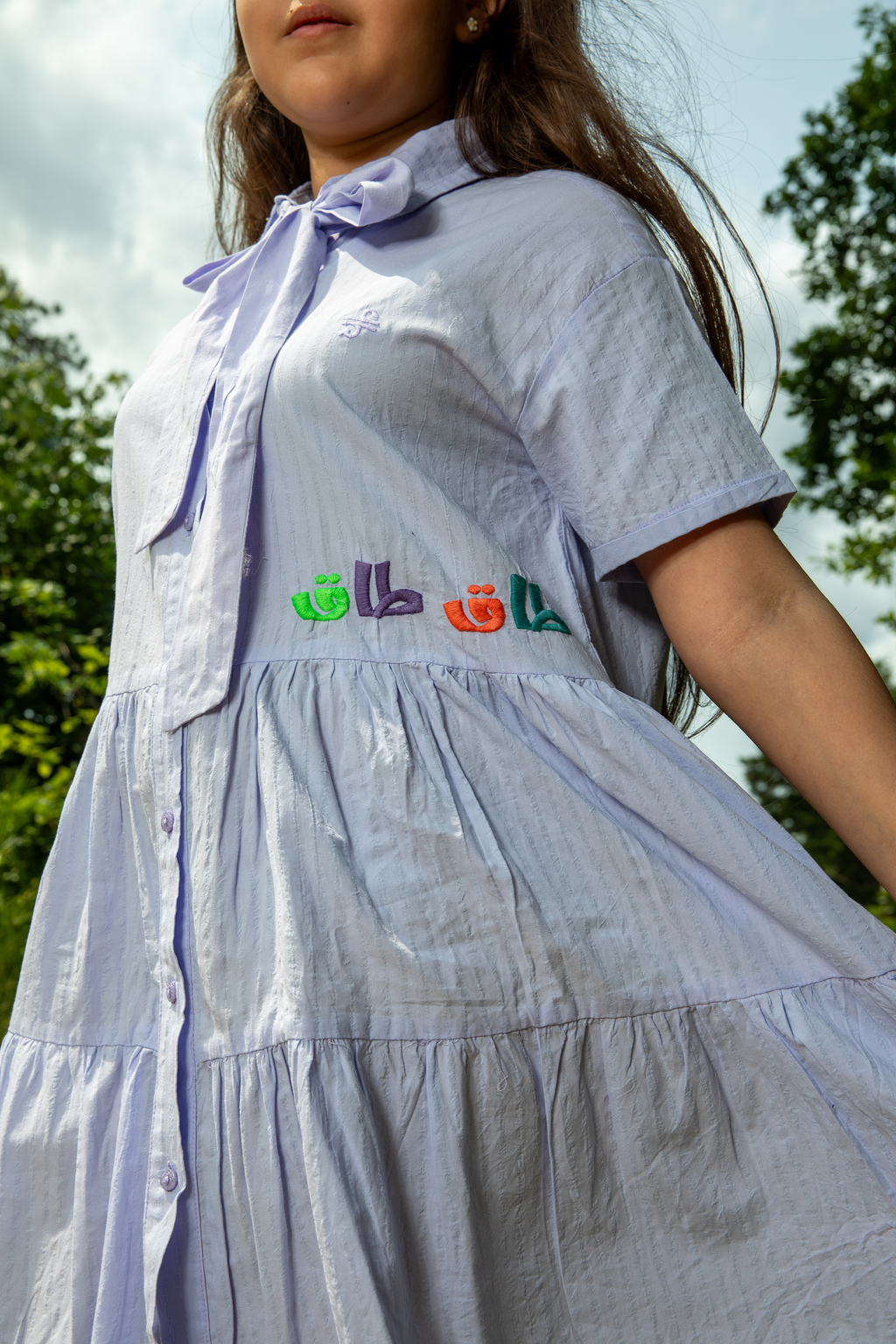 Girl wearing light purple button-down dress with embroidered colorful Arabic text outdoors