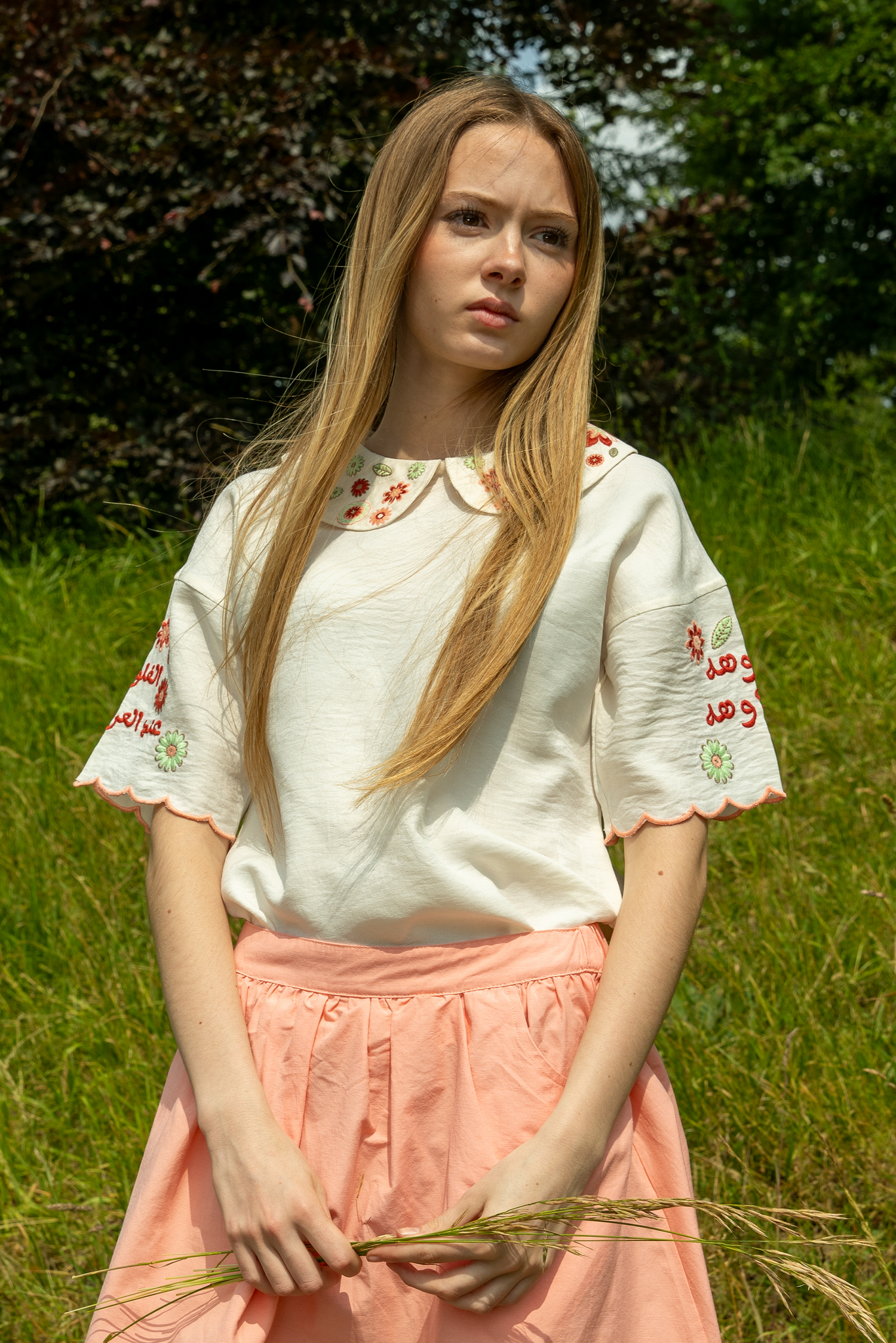 Woman in a white embroidered top and pink skirt standing in a grassy field.